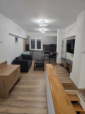 A modern living space with a wooden bench, a sofa, a dining table, and a kitchen area with cabinets and a microwave. - Modern apartment in Thessaloniki (Θεσσαλονίκη)