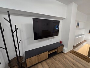 A modern living room with a flat-screen TV mounted on the wall, a wooden entertainment unit below, and a black coat rack to the left. - Modern apartment in Thessaloniki (Θεσσαλονίκη)