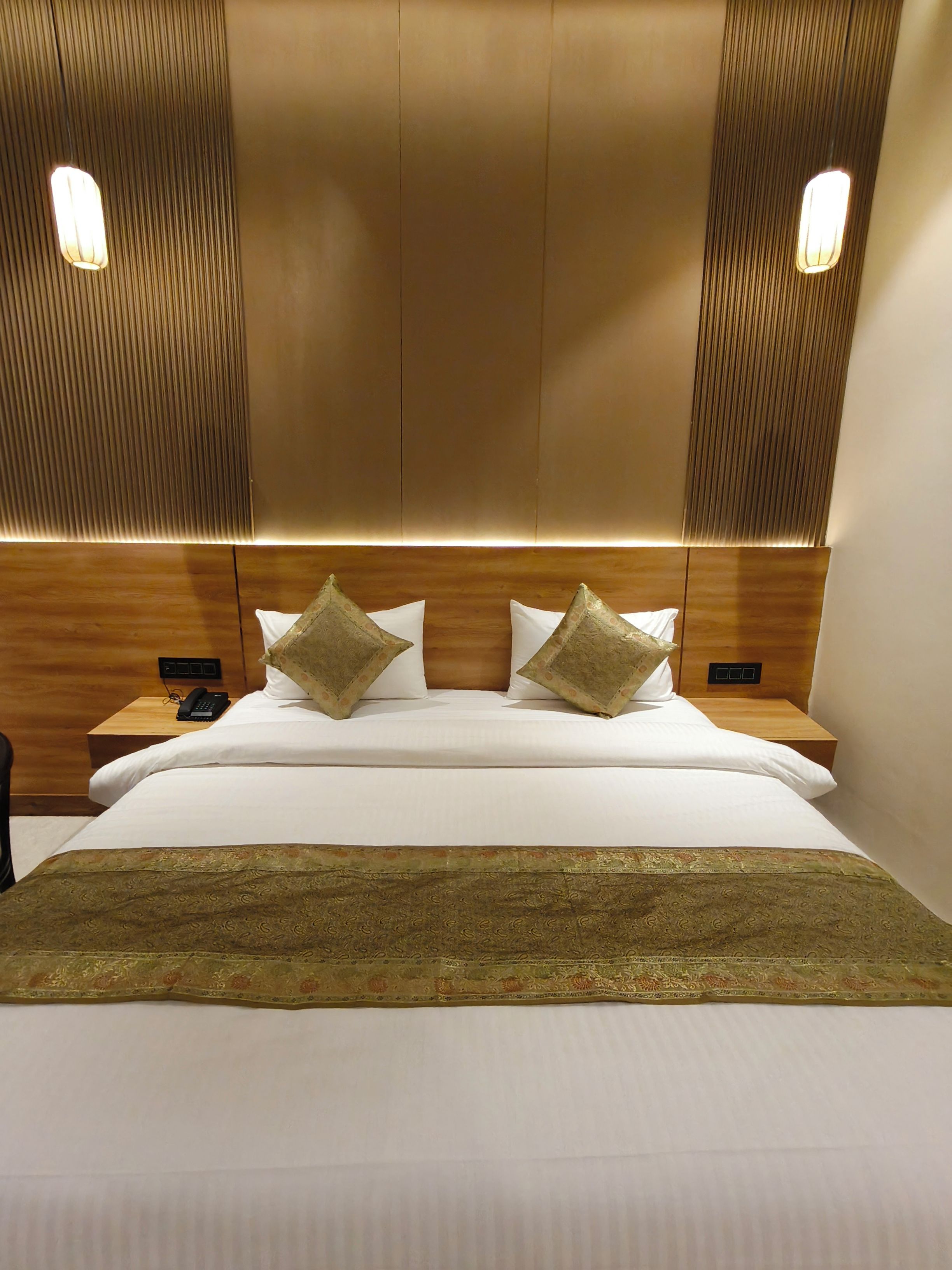A hotel room with a large bed, two bedside tables, a telephone, and a headboard with vertical lines and wall-mounted lights.