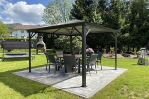 A covered patio area with a table and chairs, a stone-tiled floor, and a grill in the background. - Holiday apartment with 1 bedroom (Bodenmais)