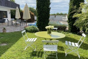Pátio com mesa redonda e cadeiras brancas, cercado por vegetação e uma piscina.