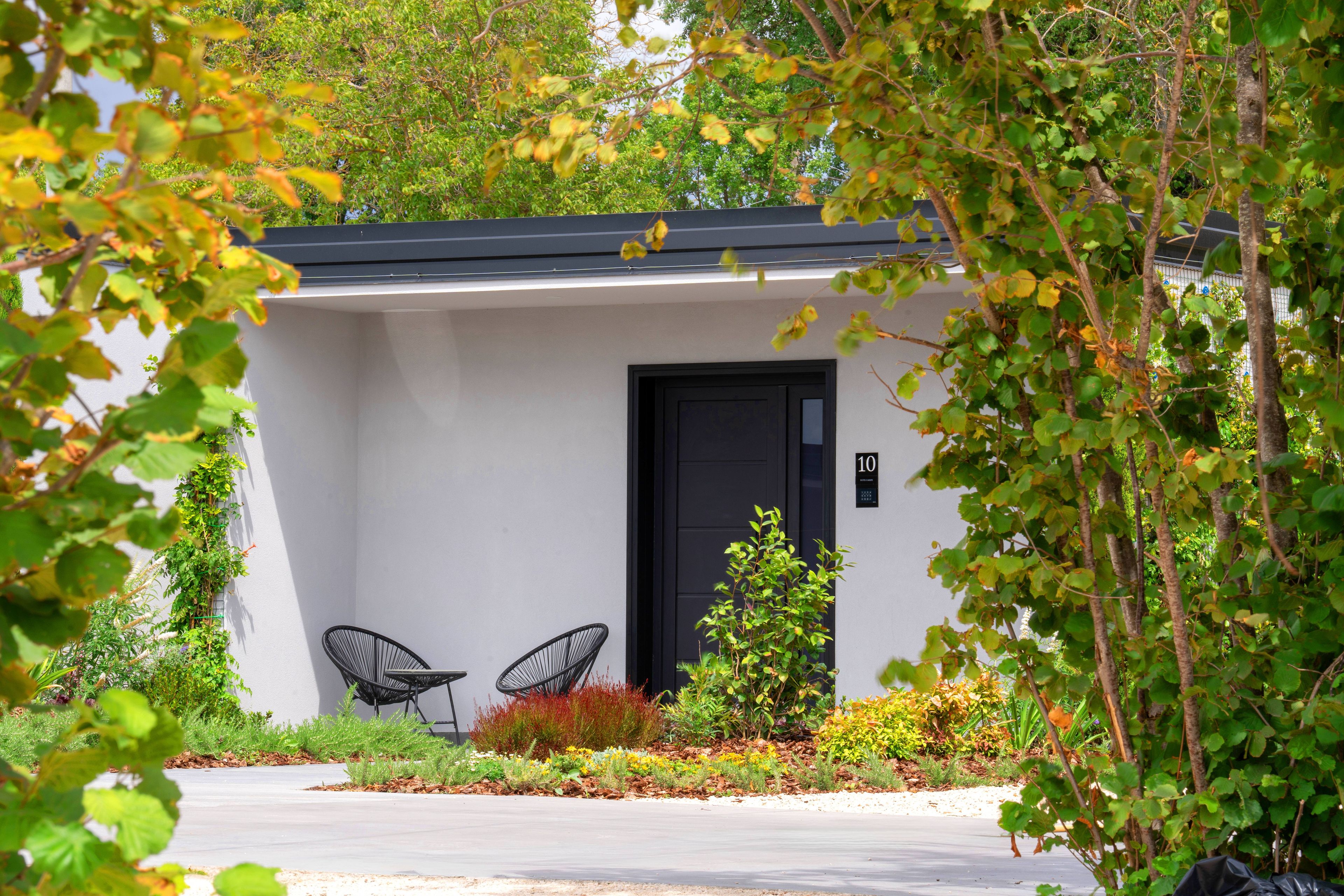 A modern building with a black door, two black chairs, and a garden with various plants.