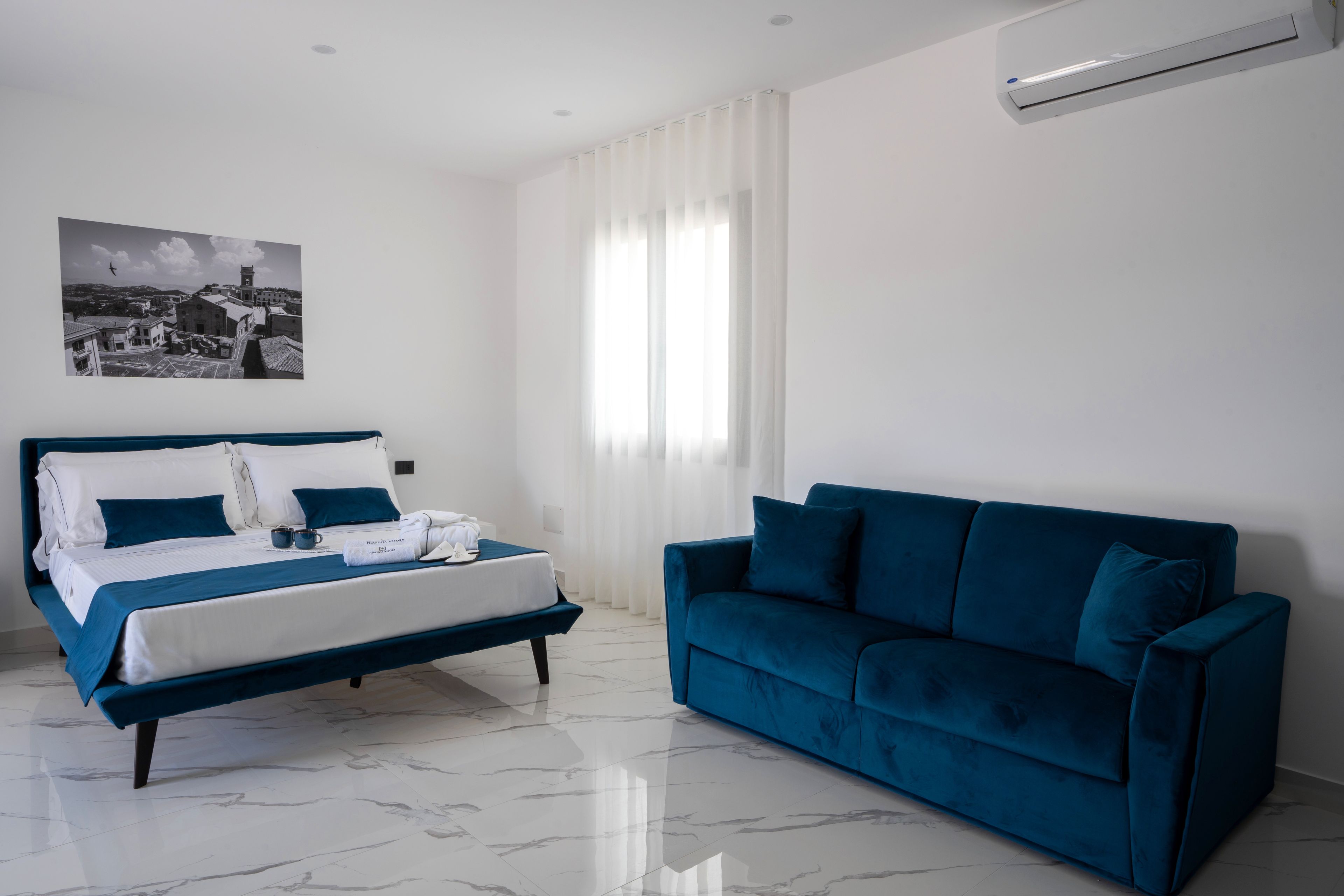 A hotel room with a bed, a blue sofa, a marble floor, a window with curtains, and a wall-mounted air conditioner.