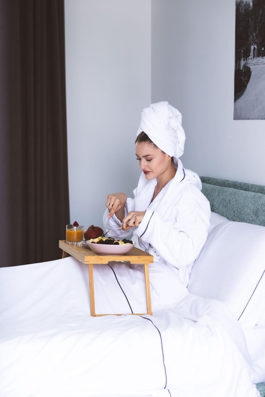 A person in a white bathrobe sitting on a bed with a tray containing a bowl of fruit and a glass of orange juice.