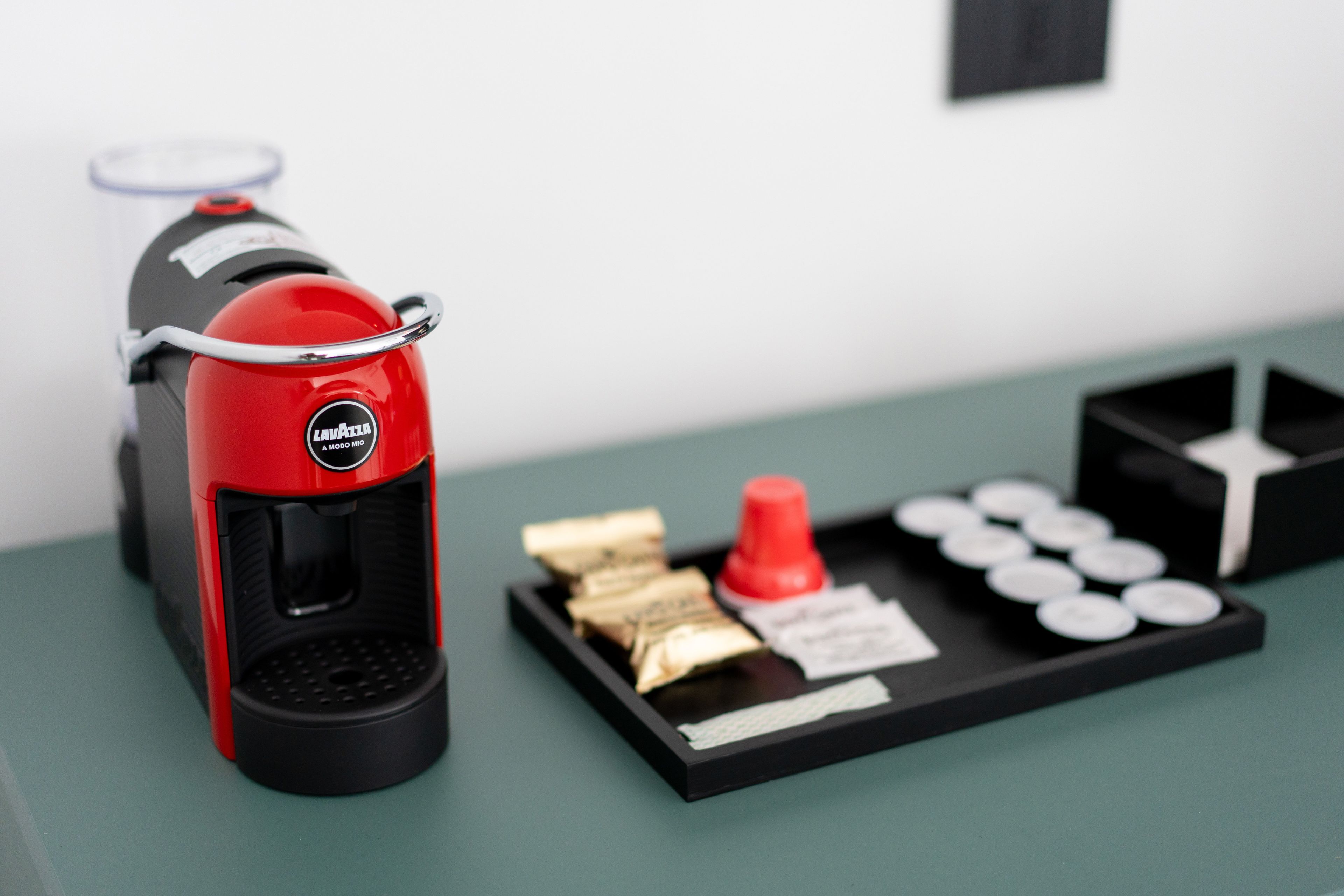 Une machine à café au design rouge et noir, un présentoir avec des capsules de café, une tasse rouge et un plateau noir avec une étiquette blanche.