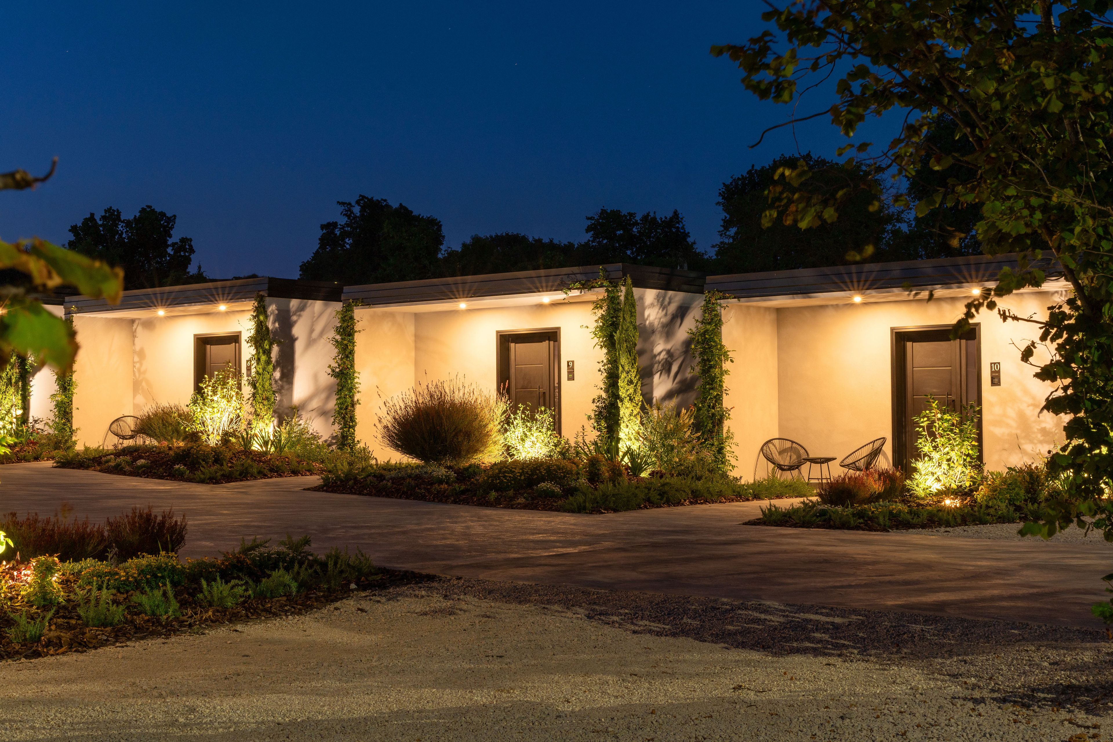 A row of modern, single-story buildings with numbered doors, surrounded by landscaped gardens and outdoor seating.