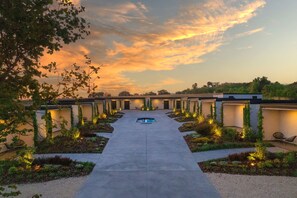 A pathway leading to a pool with a sunset sky, surrounded by greenery and illuminated lights.