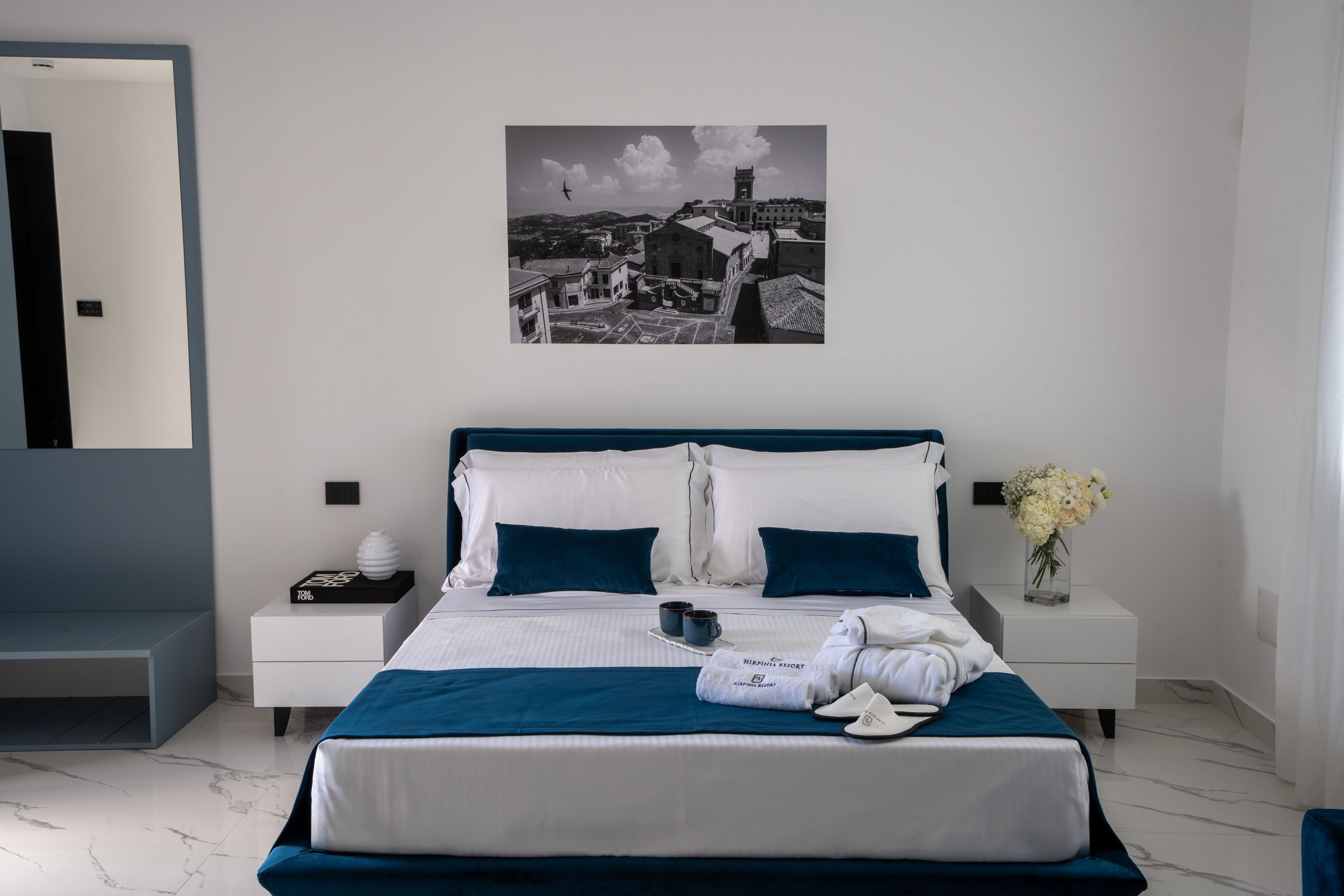 Una moderna camera d'albergo con un letto grande, un comodino, uno specchio e un dipinto raffigurante un panorama urbano.