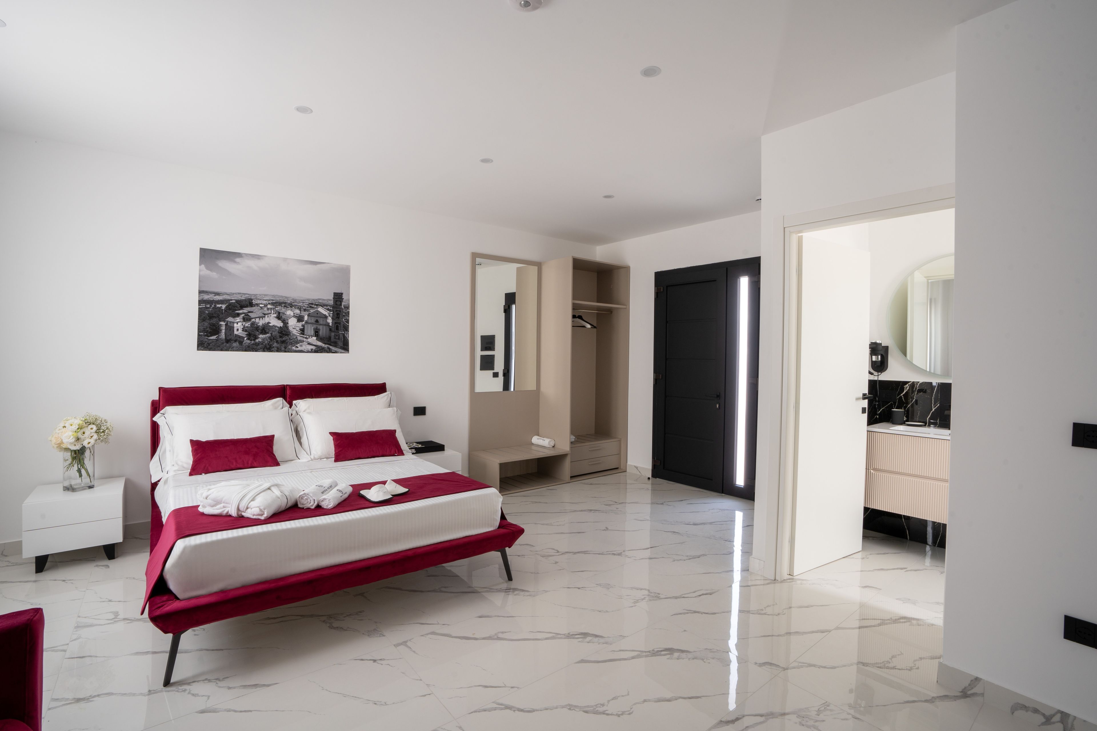 Un dormitorio moderno con cama, mesita de noche, espejo y baño visible a través de una puerta abierta.