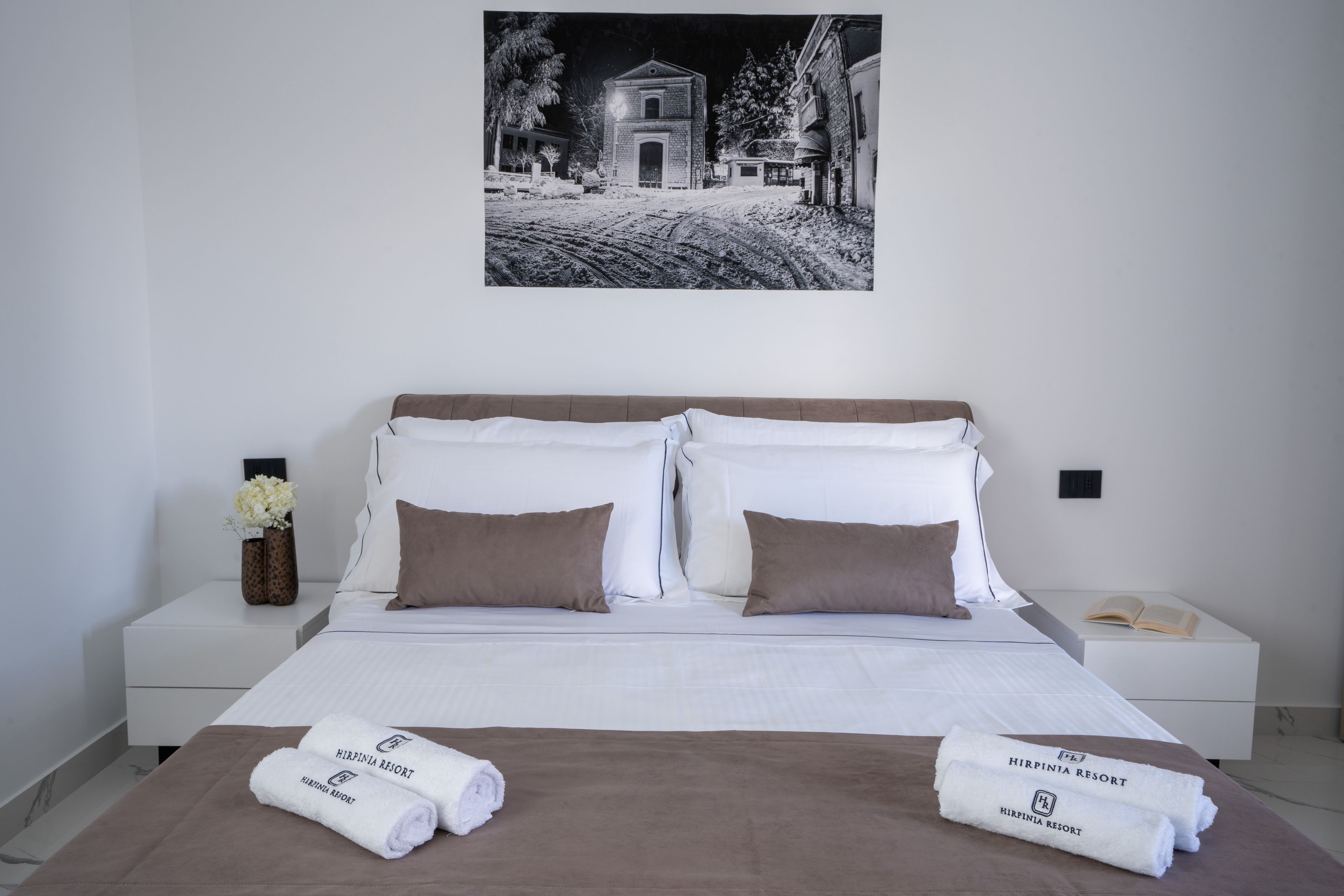 Una camera d'albergo con un letto, due cuscini, un comodino con un telefono, un vaso con fiori e un quadro appeso al muro.