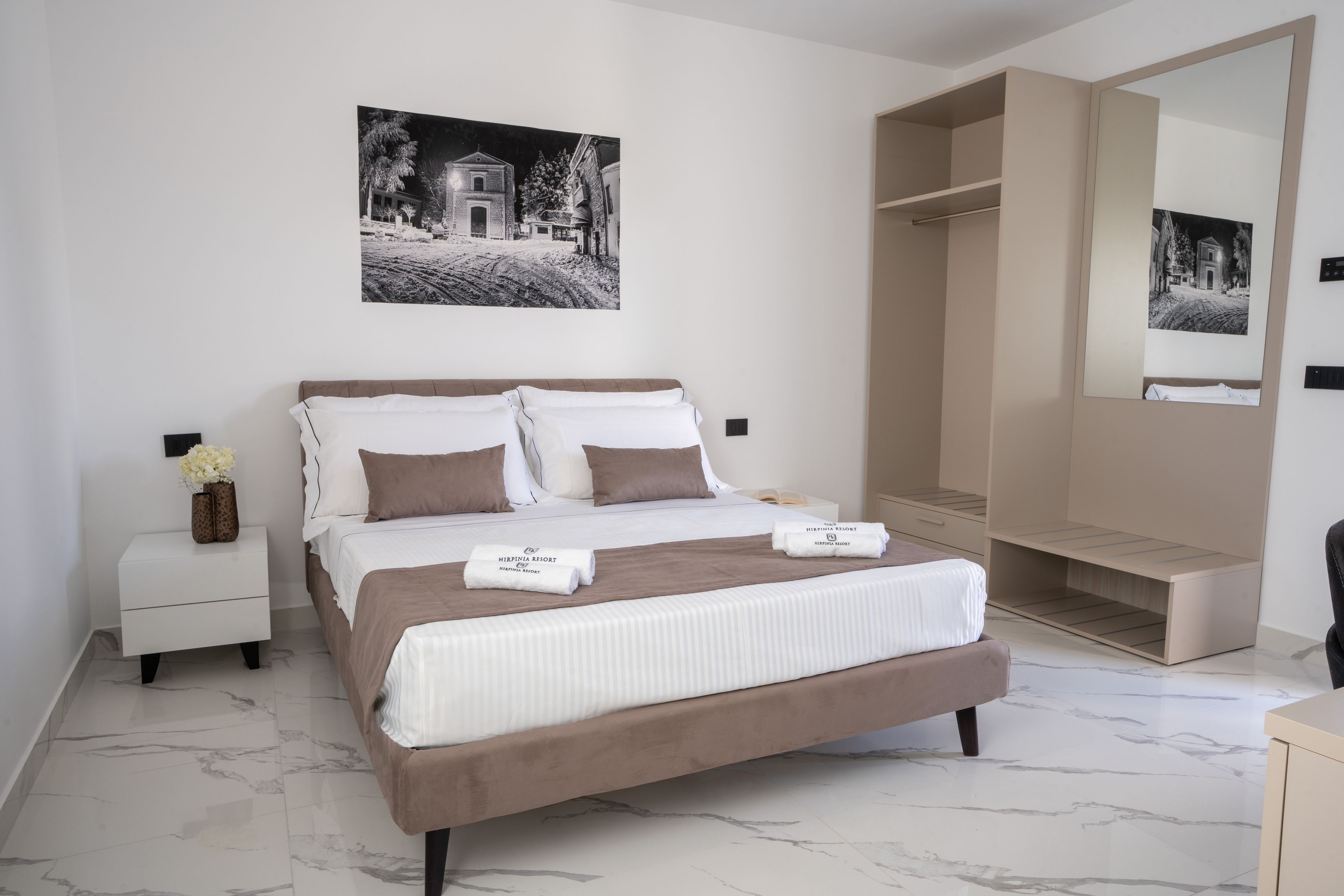 Une chambre à coucher comprenant un lit, une table de chevet, une petite table de nuit avec un vase, un grand miroir et une armoire.