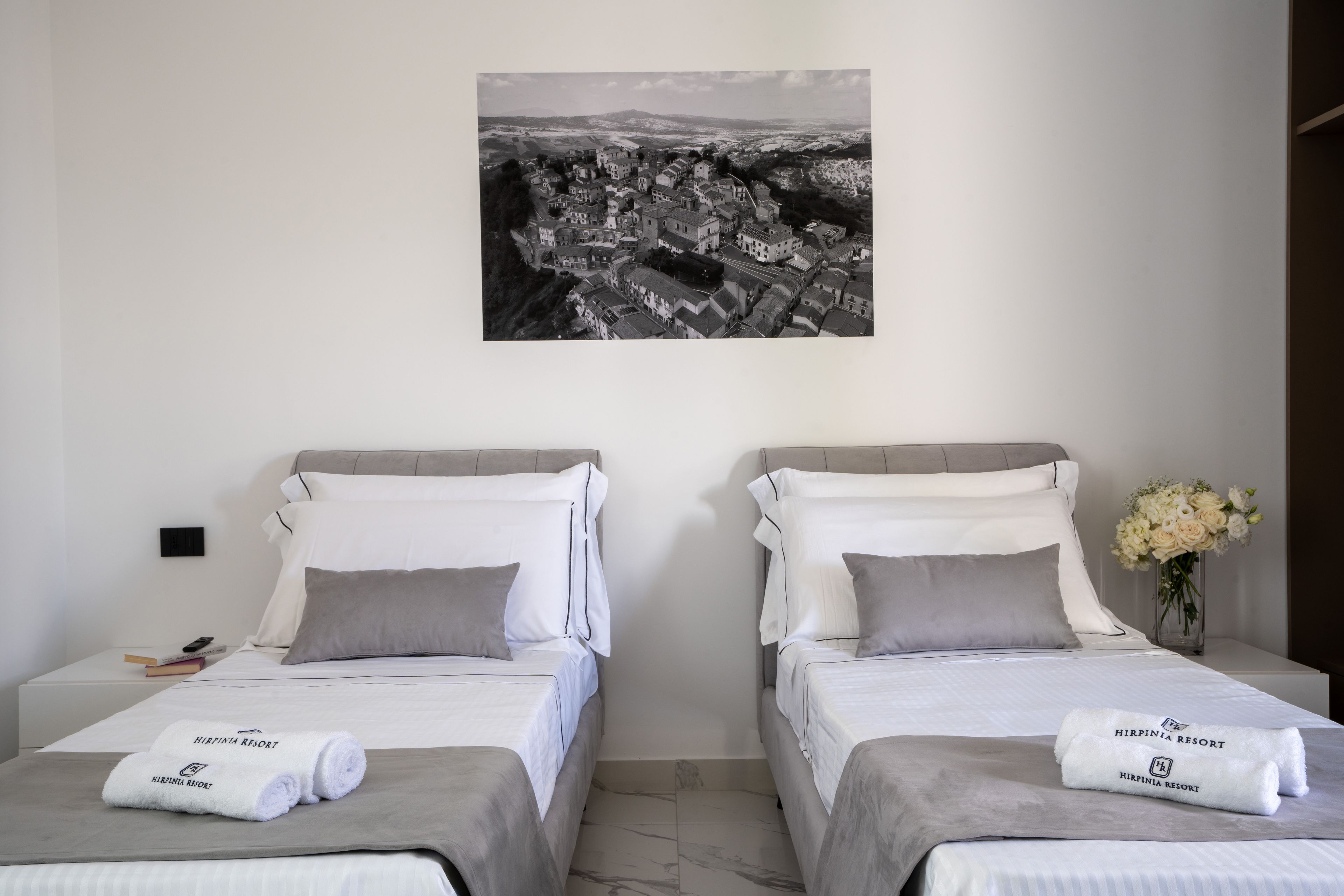 Et hotelværelse med to senge, hver med en pude og et sammenrullet håndklæde. Der er et indrammet fotografi på væggen over sengene.