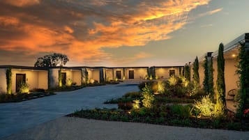 Un cortile con un sentiero che conduce a un edificio con diverse porte, circondato da vegetazione e illuminato da luci soffuse.
