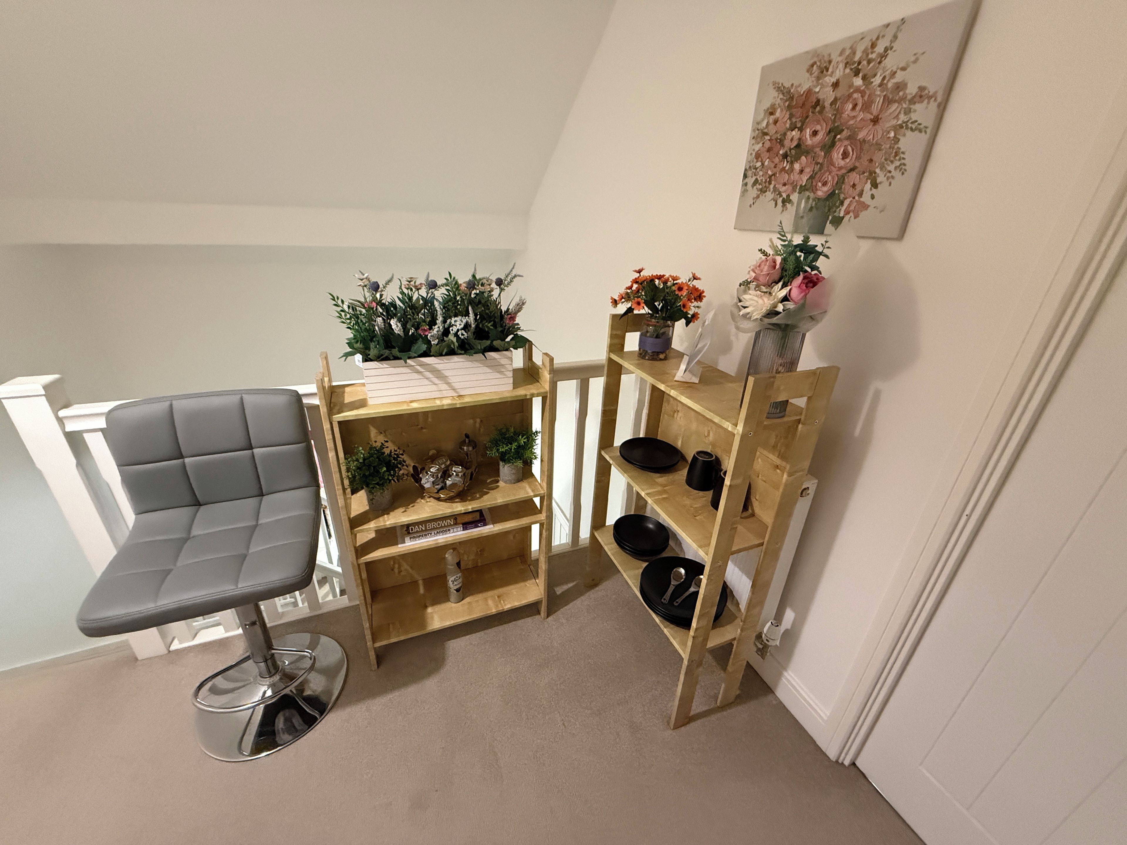 Une pièce d’angle avec un fauteuil à revêtement gris, une étagère en bois ornée de plantes et d’objets décoratifs, et un tableau accroché au mur.