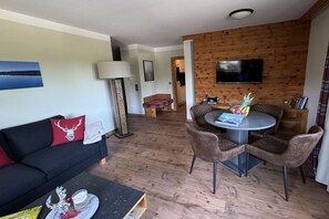 Un salon avec un canapé noir, une table ronde entourée de chaises, un téléviseur fixé à un mur en bois et une lampe.