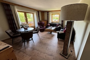 A room with a wooden floor, a round table with chairs, a sofa, a lamp, and a window with curtains. - Holiday apartment with 1 bedroom (Schwangau)