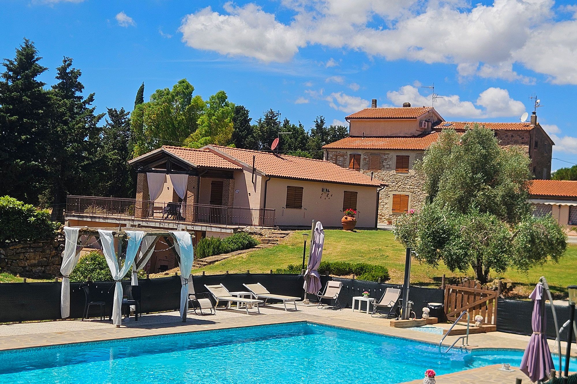 Una piscina con sillones de descanso y sombrillas, rodeada por un edificio de piedra con techo de tejas.