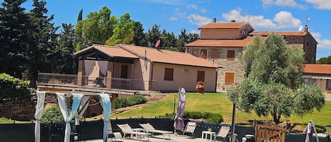 Una piscina con sillones de descanso y sombrillas, rodeada por un edificio de piedra con techo de tejas.