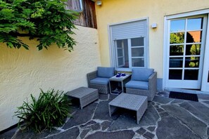 An outdoor seating area with wicker furniture, a stone floor, and a window with a closed shutter. - Holiday apartment with 1 bedroom (Endingen-Königschaffhausen)