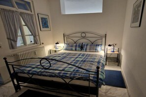 A bedroom with a metal bed frame, a checkered bedspread, a window with curtains, a bedside table with a lamp, and a radiator. - Holiday apartment with 1 bedroom (Endingen-Königschaffhausen)