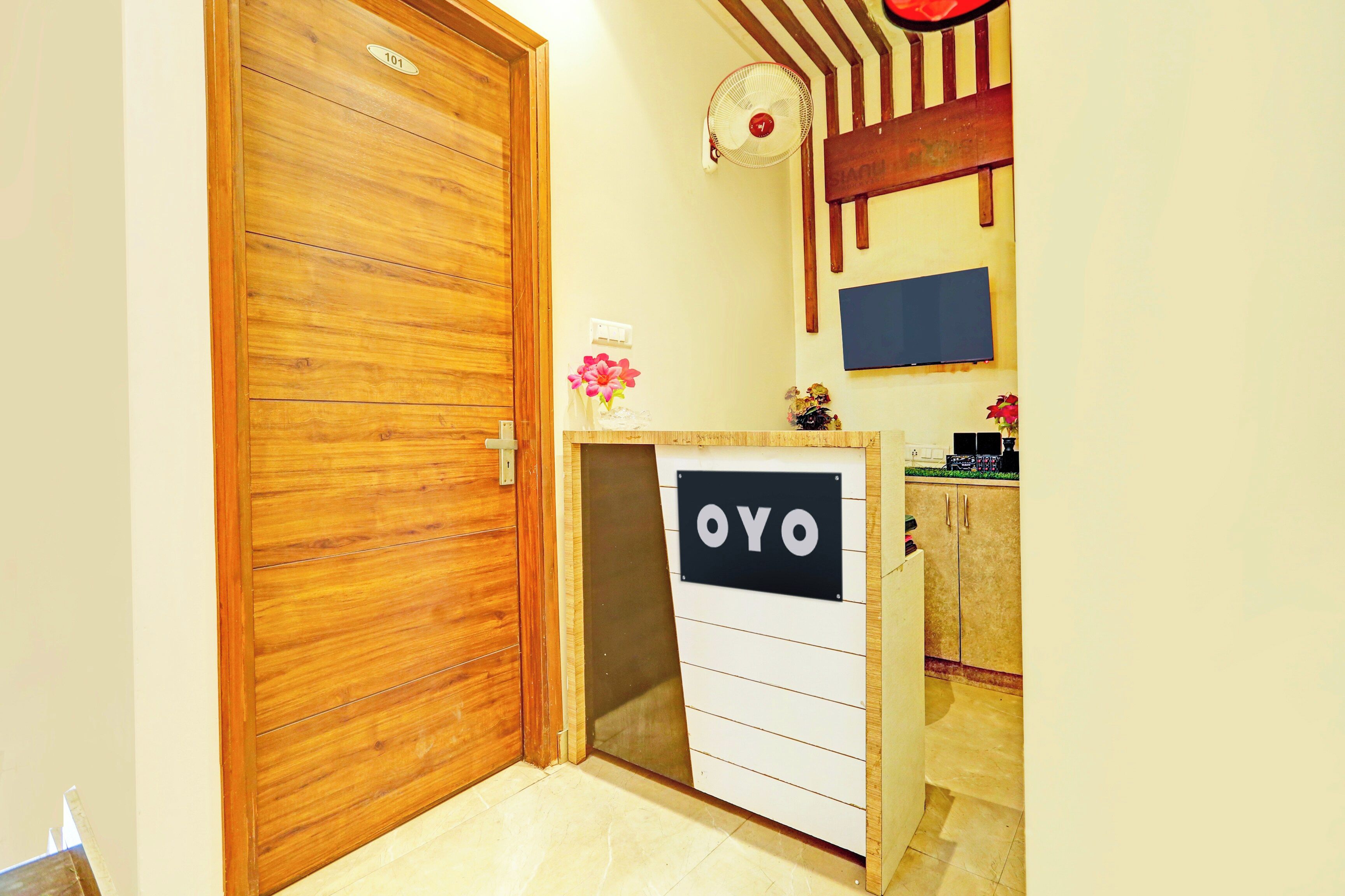 A room with a wooden door, a small desk with a sign that reads 'OYO', a wall-mounted fan, and a television.