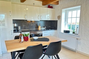 A modern kitchen with white cabinets, a wooden dining table, and a window with a view. - Holiday home with 2 bedrooms (Büsumer-Deichhausen)