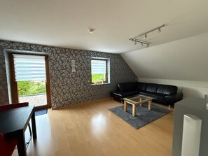 En moderne stue med en sort lædersofa, et træsofabord og et spisebord med stole. To vinduer med persienner, en loftlampe og en væg med mønster.