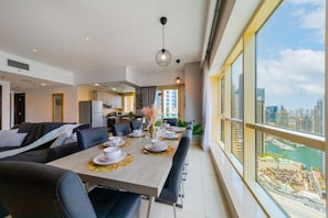 A modern dining area with a long table set for four, a kitchen in the background, and a view of a marina through large windows. - Royal Oceanic 2BR with Marina View (Dubai)