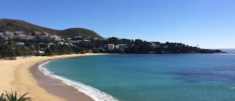 Un paisaje costero con playa arenosa, aguas azules cristalinas y una colina con edificios.