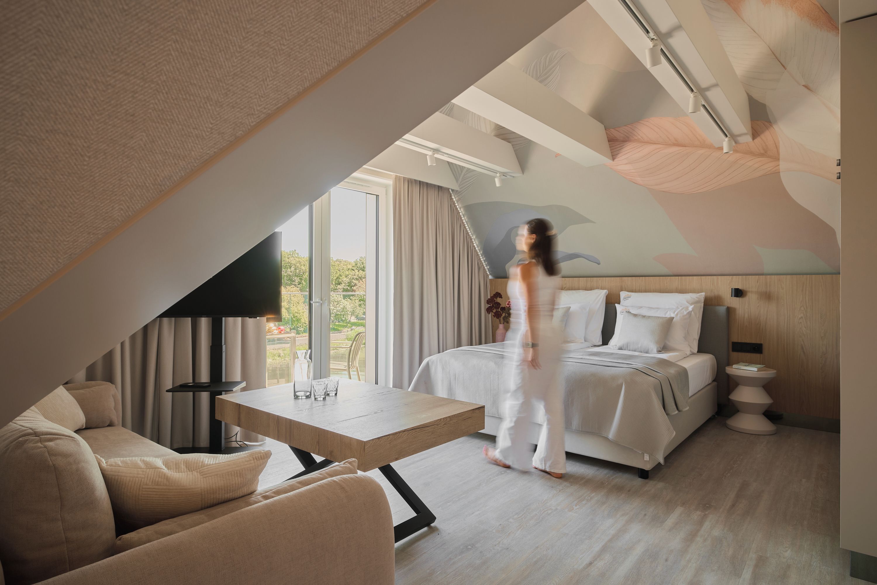 A modern hotel room with a large bed, a sitting area with a table, and a view of the outdoors.