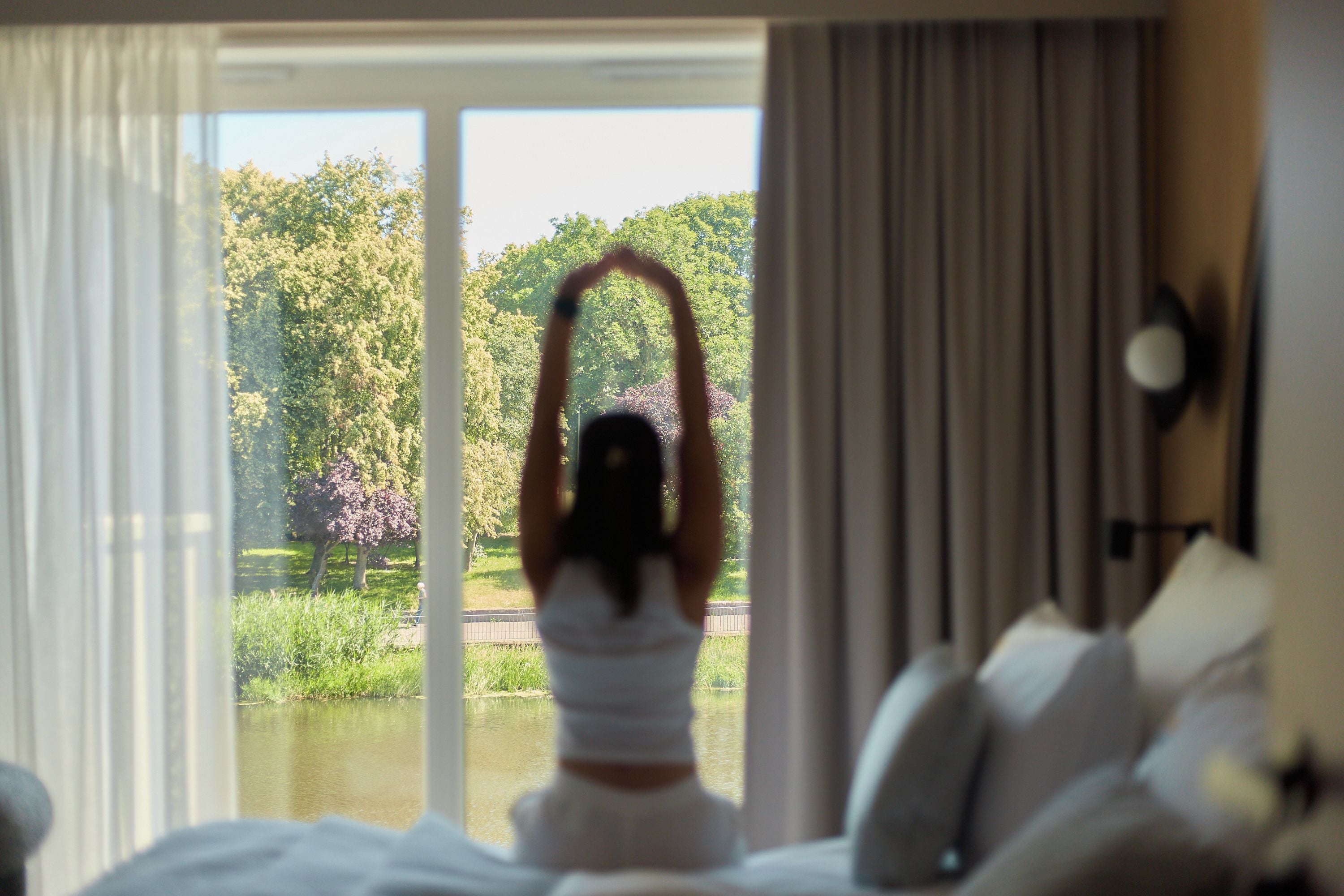 Ein Zimmer mit großem Fenster, einem Bett und Blick auf einen Park mit Bäumen und einem Gewässer.