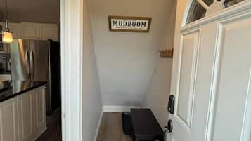 A narrow hallway leading to a kitchen with white cabinetry and a black countertop. A small table with a chair is placed against the wall, and a sign above reads 'MUDROOM.'