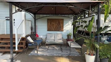 A covered patio area with outdoor furniture, a patterned rug, and potted plants.