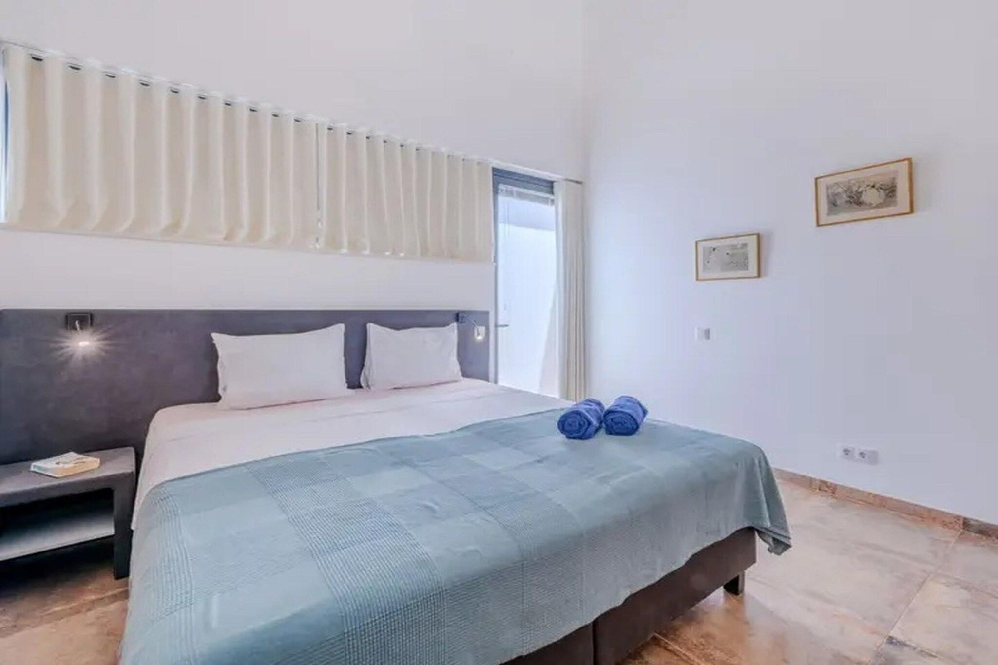 A hotel room with a bed, a bedside table, a window with curtains, and two wall-mounted pictures.