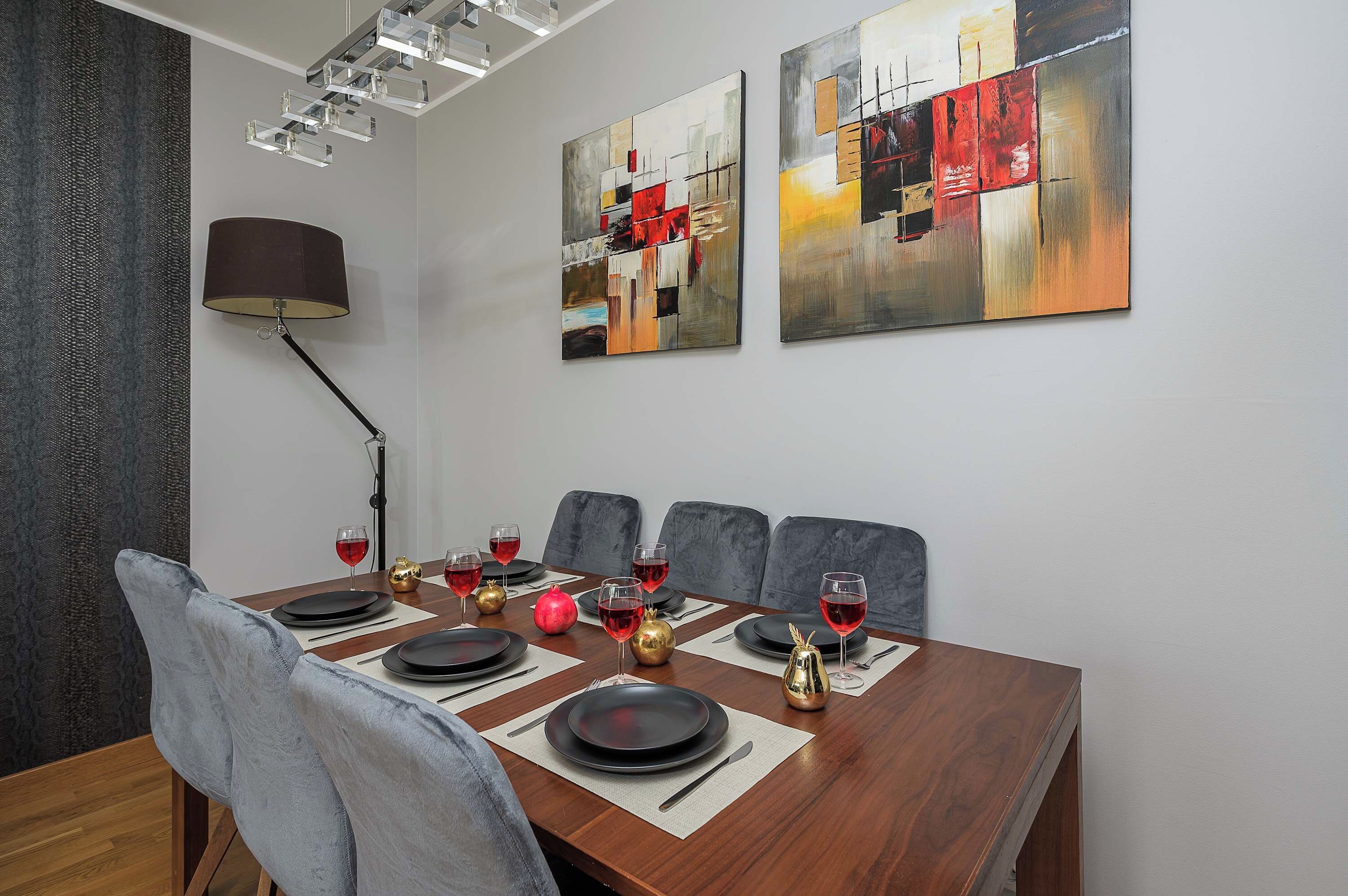 Una sala da pranzo con un tavolo in legno apparecchiato per sei persone, sedie rivestite in grigio e due dipinti astratti appesi al muro.