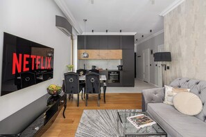 A modern living room with a flat-screen TV, a sofa, a coffee table, and a dining area with chairs and a kitchen in the background. - Lumina 3-room Apart w/ Terrace I Parking (Warszawa)