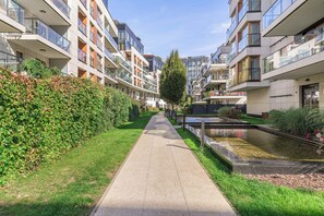 A pathway flanked by modern apartment buildings with balconies, a grassy area, and a small water feature. - Lumina 3-room Apart w/ Terrace I Parking (Warszawa)