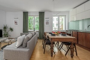 A modern living room with a grey sofa, dining table with chairs, and a kitchen area with a stove and cabinets. - Lumina Comfy 2BR Apart near the Center of Warsaw (Warszawa)