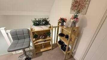 A corner room with a grey upholstered chair, a wooden shelf unit with plants and decorative items, and a painting on the wall.