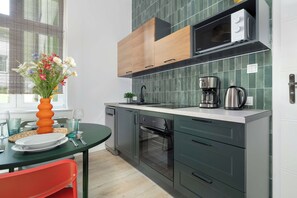 A well-equipped kitchenette displays dark green lower cabinetry paired with light wooden upper shelves and countertops. A small dining area includes a dark table surrounded by vibrant orange and red chairs. - Poznan Dabrowskiego 49 Spacious Apartment Balcony (Poznań)
