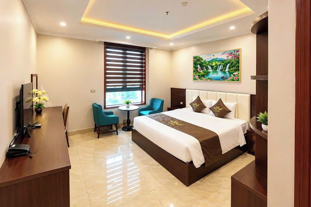 A hotel room with a large bed, a desk with a computer, a chair, a window with blinds, and a painting on the wall.