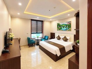 A hotel room with a large bed, a desk with a computer, a chair, a window with blinds, and a painting on the wall. - MAISON PARK (Vo Cuong)
