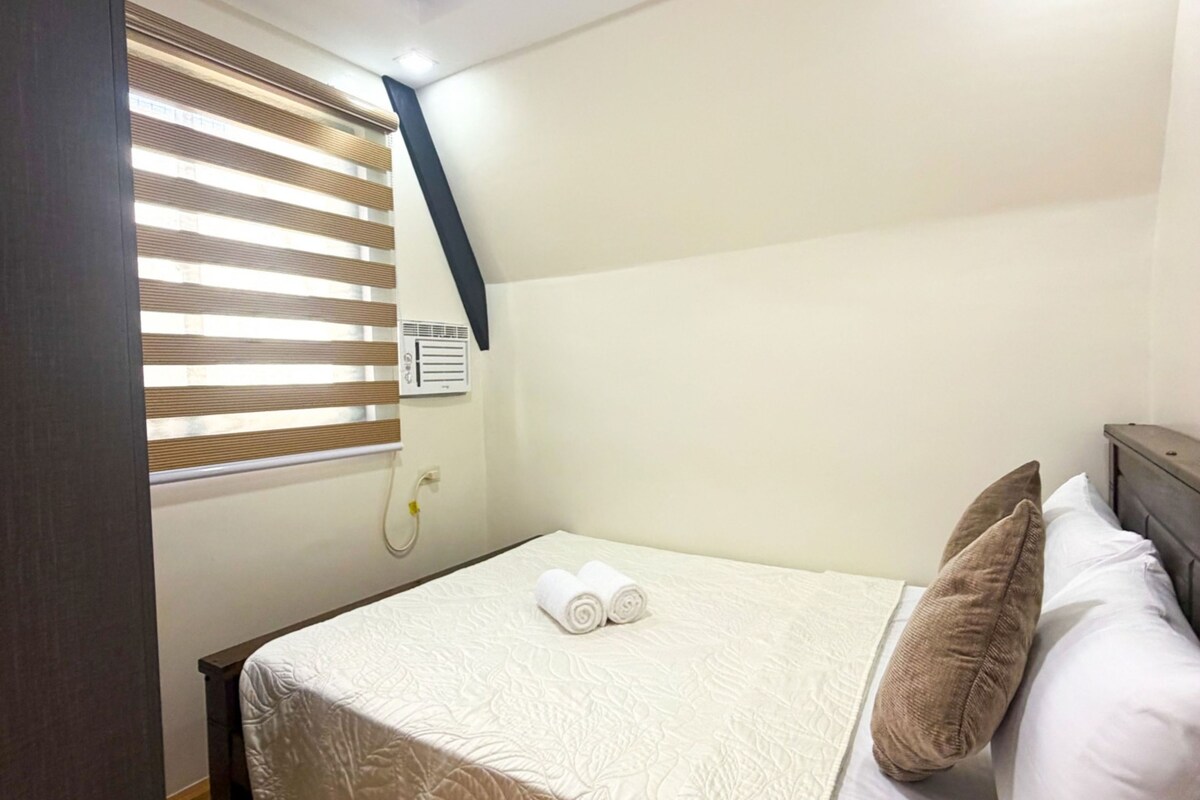 Een hotelkamer met een bed, kussens, een raam met jaloezieën en een wandradiator.