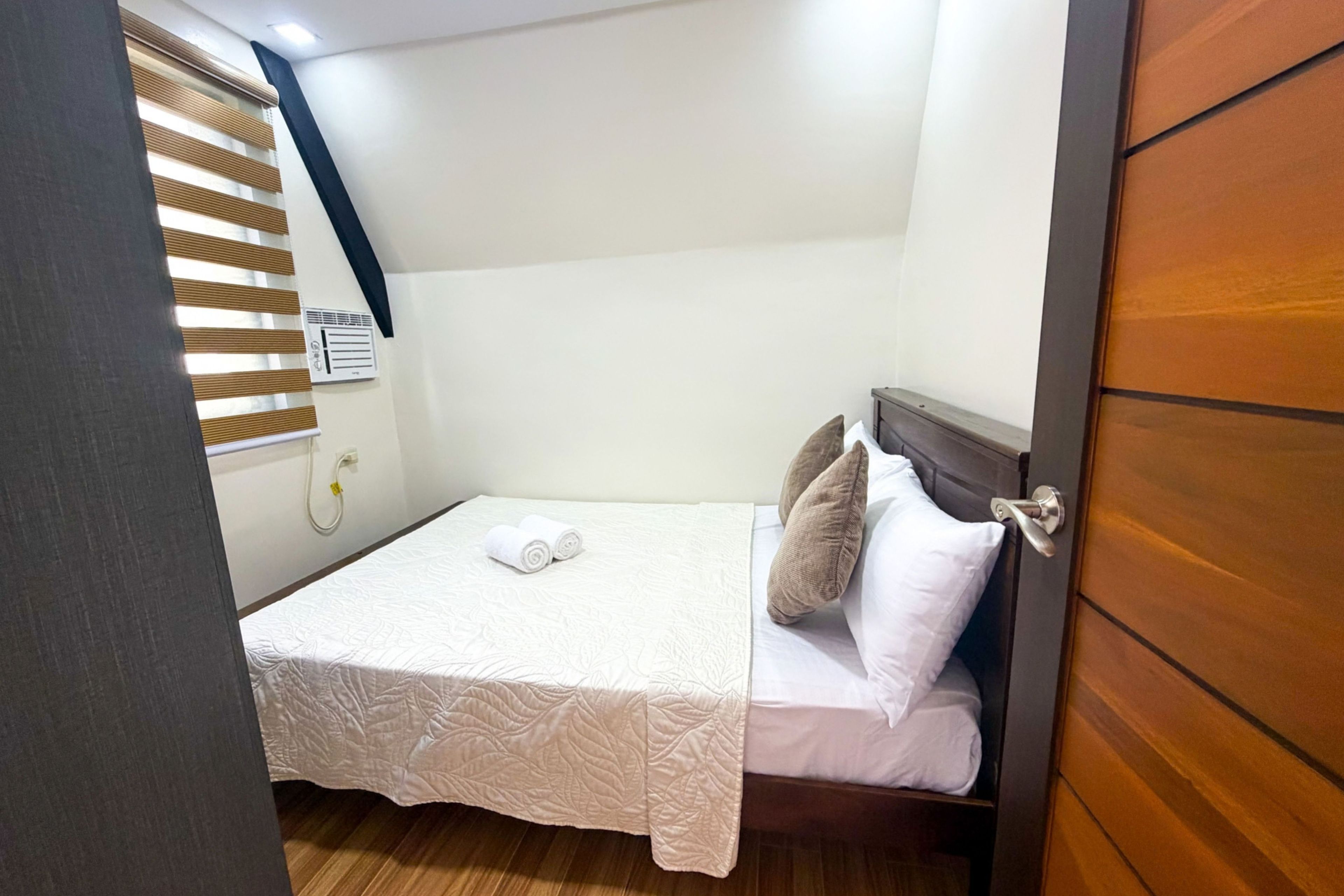 Una cama pequeña y bien hecha con colcha y almohadas blancas, un cabecero de madera y una ventana con persianas.