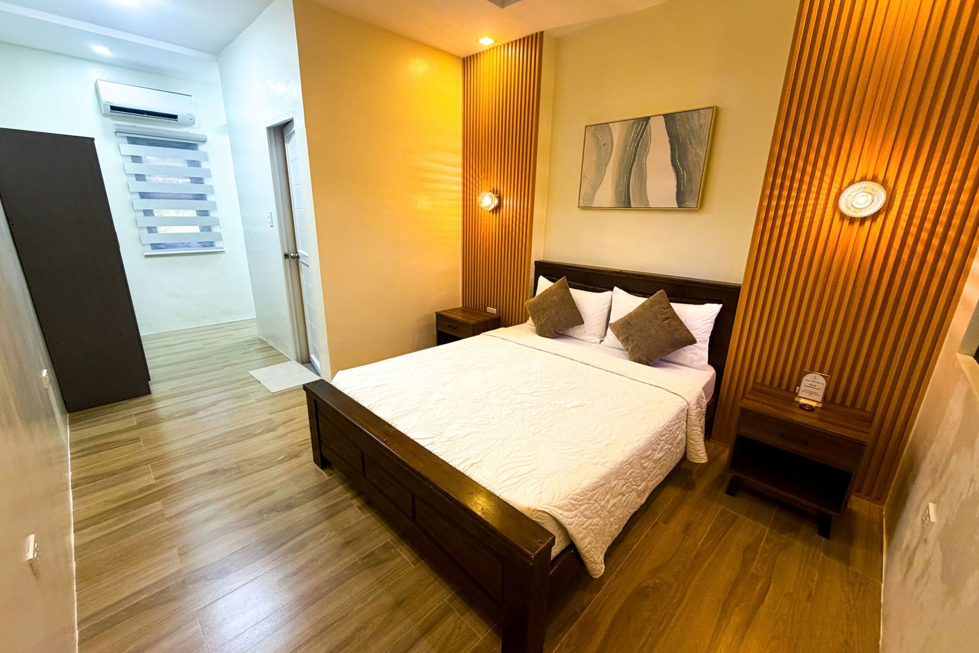 A hotel room with a wooden bed, a nightstand, a wall-mounted light, and a painting.