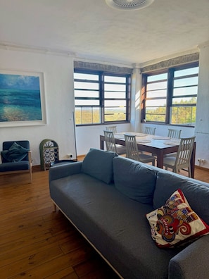A living room with a gray sofa, a dining table with chairs, and large windows with a view. - Secluded Mountainside Retreat w/ Pool, Sauna + Gym (Caldas de Monchique)