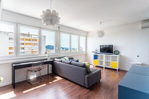 A modern living room with a sofa, a desk, and a TV. Large windows offer a view of buildings outside. - Comfortable apartment with great city and sea view (Split)