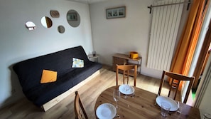 A cozy room with a black sofa, a wooden dining table with white plates and glasses, a small side table, and a window with curtains. - Studio with balcony and ski slopes view (Les Deux Alpes)