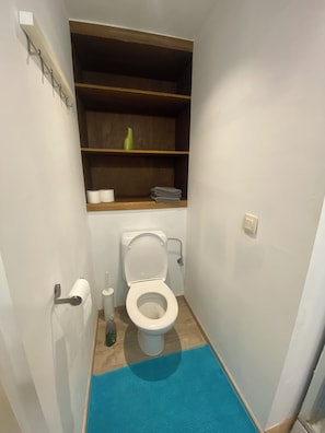 A small bathroom with a toilet, a shelf above the toilet, and a blue mat on the floor. - Studio with air conditioning and bike storage (Huy)