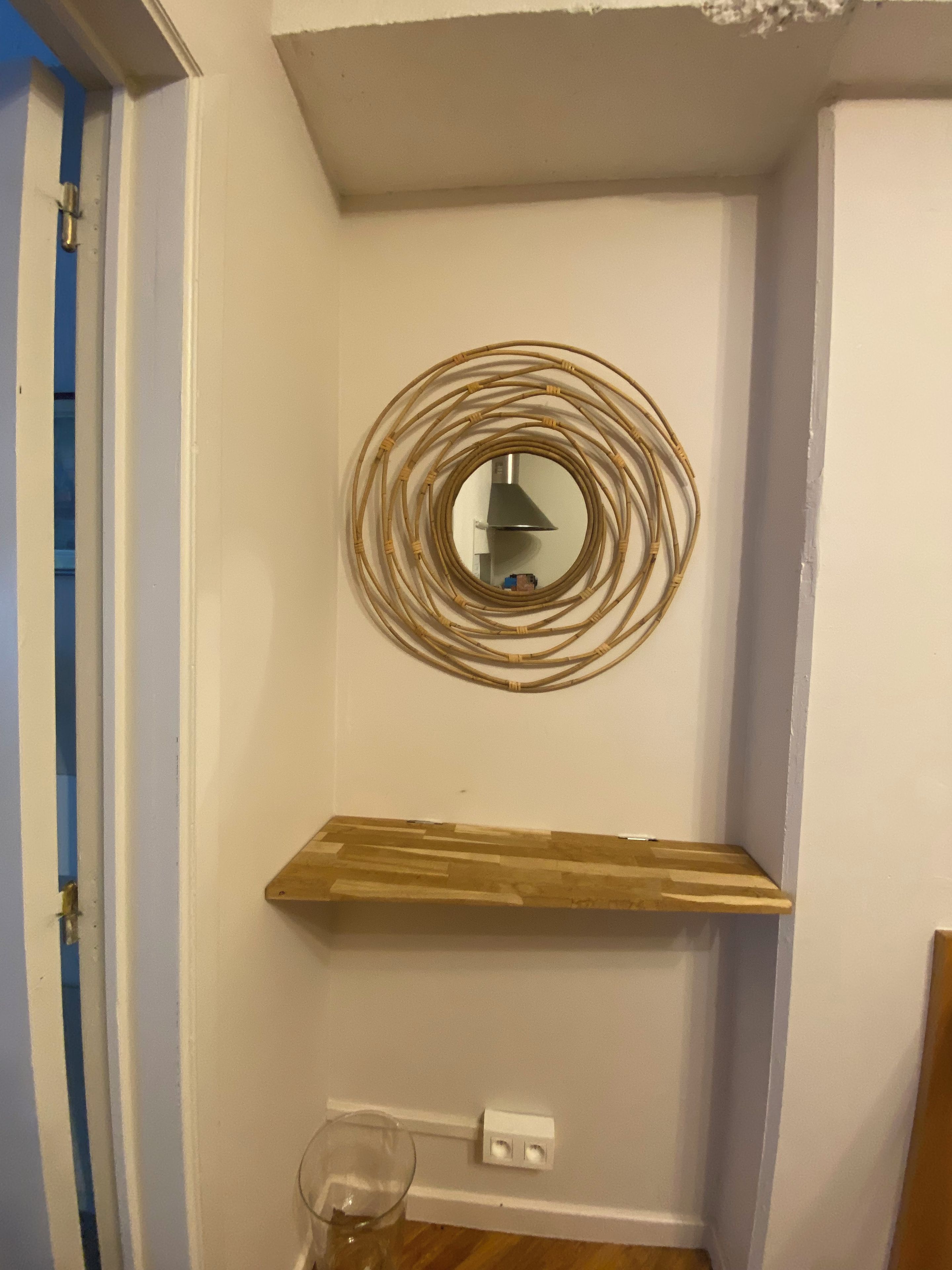 Een hoek van een kamer met een wandspiegel, een houten plank en een stopcontact.
